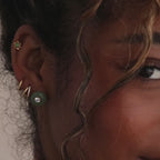 Model showcasing a gold flower-shaped jade stud earring in her cartilage, stacked with a round jade earring, and multiple gold huggie hoops.