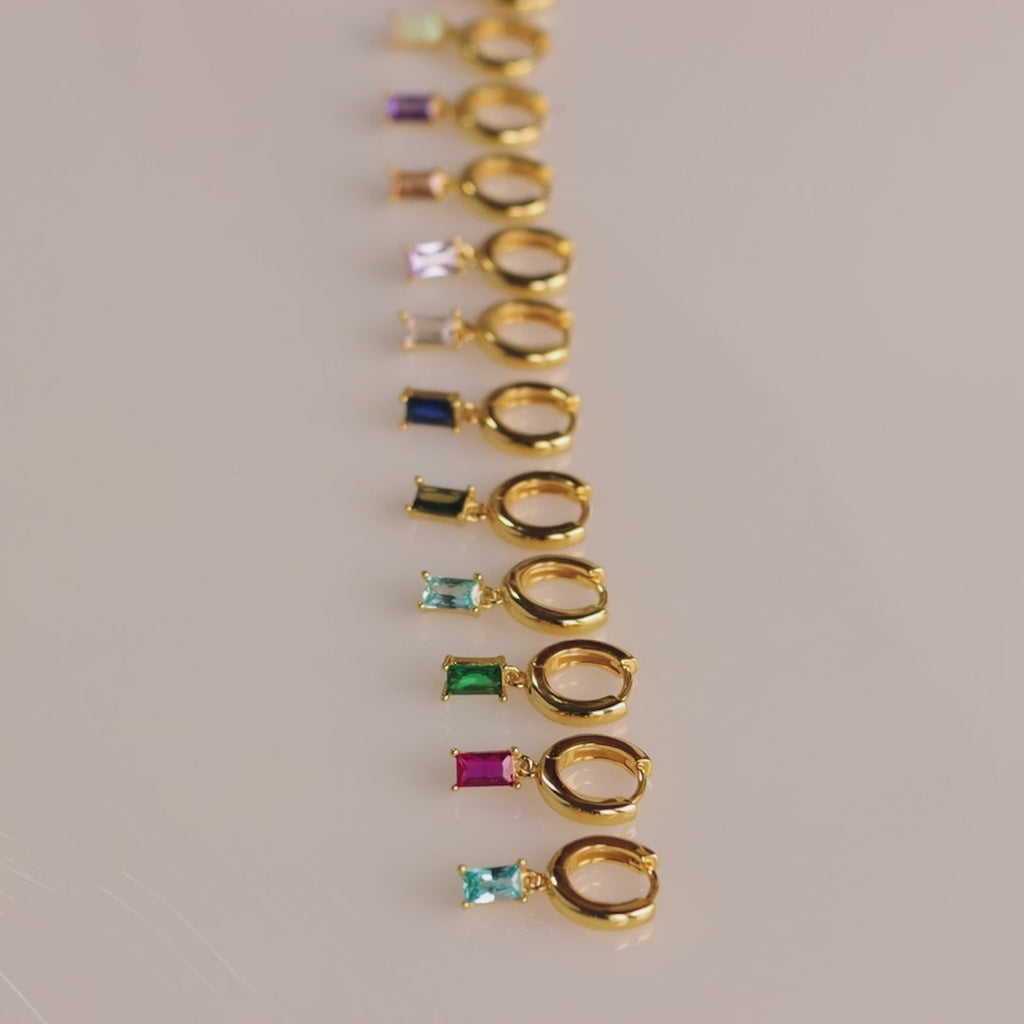 A full set of gold huggie earrings with baguette-cut birthstones arranged in a line, including garnet, sapphire, emerald, and more.