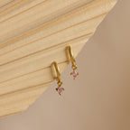 Ruby Drop Huggies, dainty gold hoop earrings with small pink gemstone charms, are displayed on a beige textured surface.