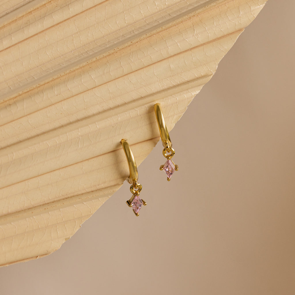 Pink Tourmaline Drop Huggies with small, pink, diamond-shaped pendants are displayed on a textured beige surface.