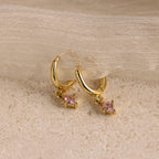Pink Tourmaline Drop Huggies, shown on a textured beige surface with sheer fabric—ideal as personalized jewelry or birthstone earrings featuring small pink gemstones.