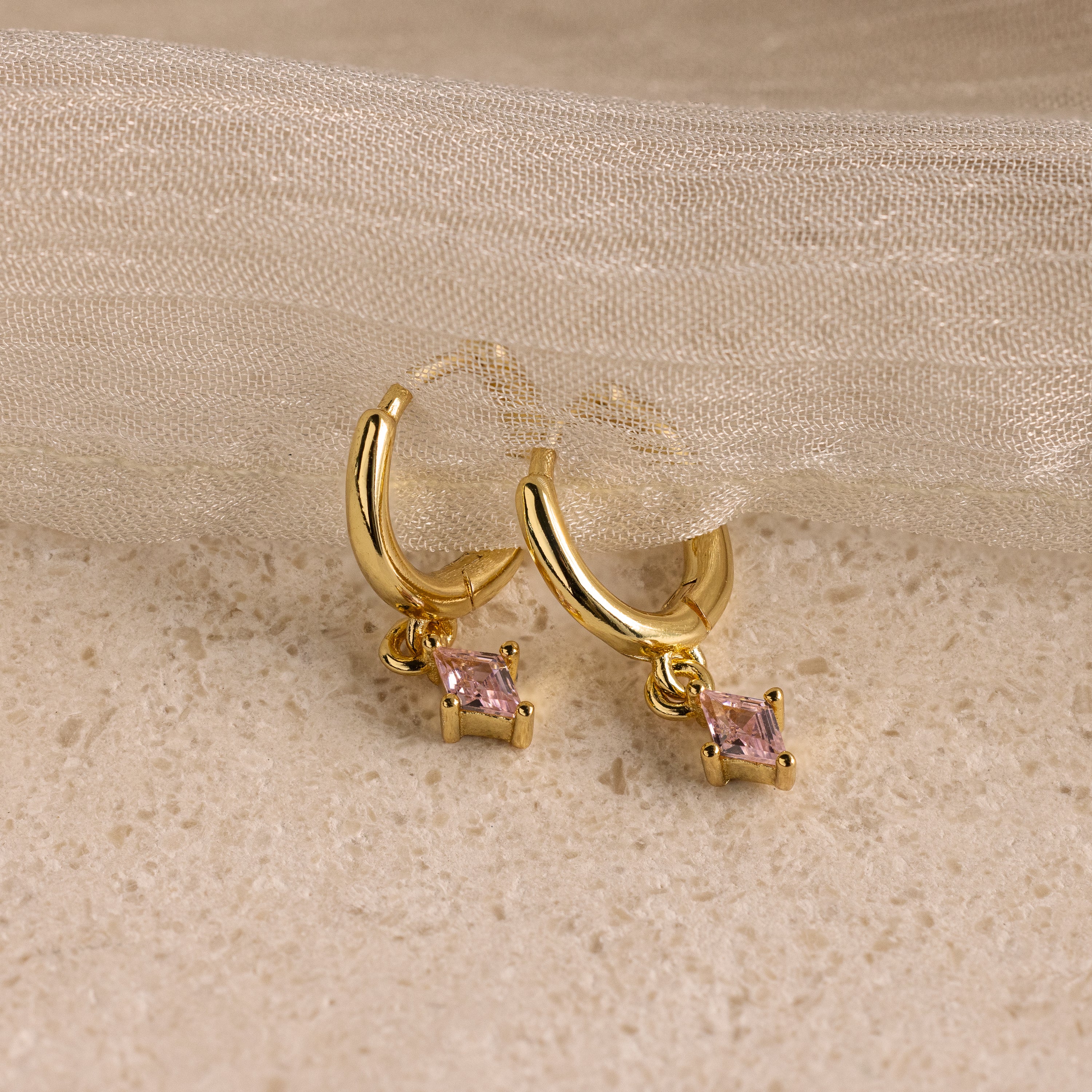 Pink Tourmaline Drop Huggies, shown on a textured beige surface with sheer fabric—ideal as personalized jewelry or birthstone earrings featuring small pink gemstones.