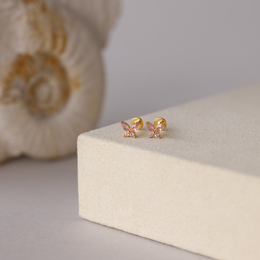 Butterfly Pink Tourmaline Flatback Studs with gold finish and pink stones shown on beige, seashell in background.