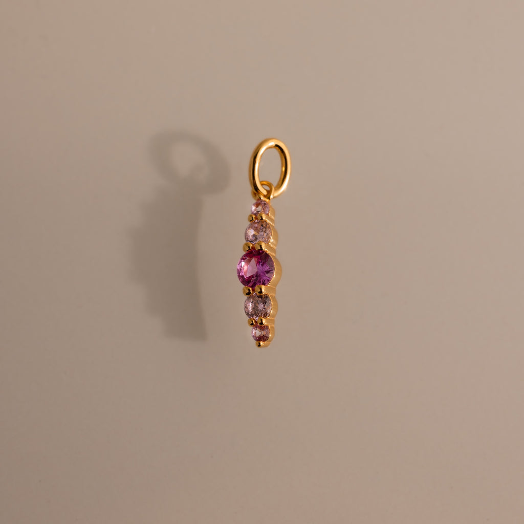 Gold pendant featuring pink tourmaline and clear stones in a vertical design, casting a shadow on beige—an elegant Ombre Lace Pink Tourmaline Charm, ideal as a personalized or birth month gift.