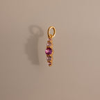 Gold pendant featuring pink tourmaline and clear stones in a vertical design, casting a shadow on beige—an elegant Ombre Lace Pink Tourmaline Charm, ideal as a personalized or birth month gift.