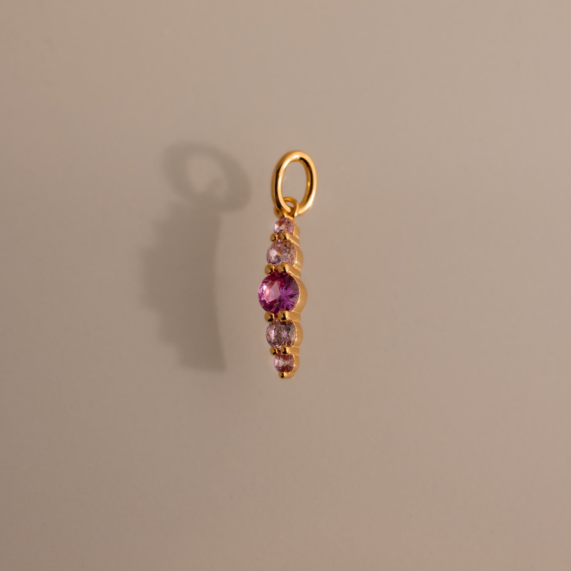 Gold pendant featuring pink tourmaline and clear stones in a vertical design, casting a shadow on beige—an elegant Ombre Lace Pink Tourmaline Charm, ideal as a personalized or birth month gift.