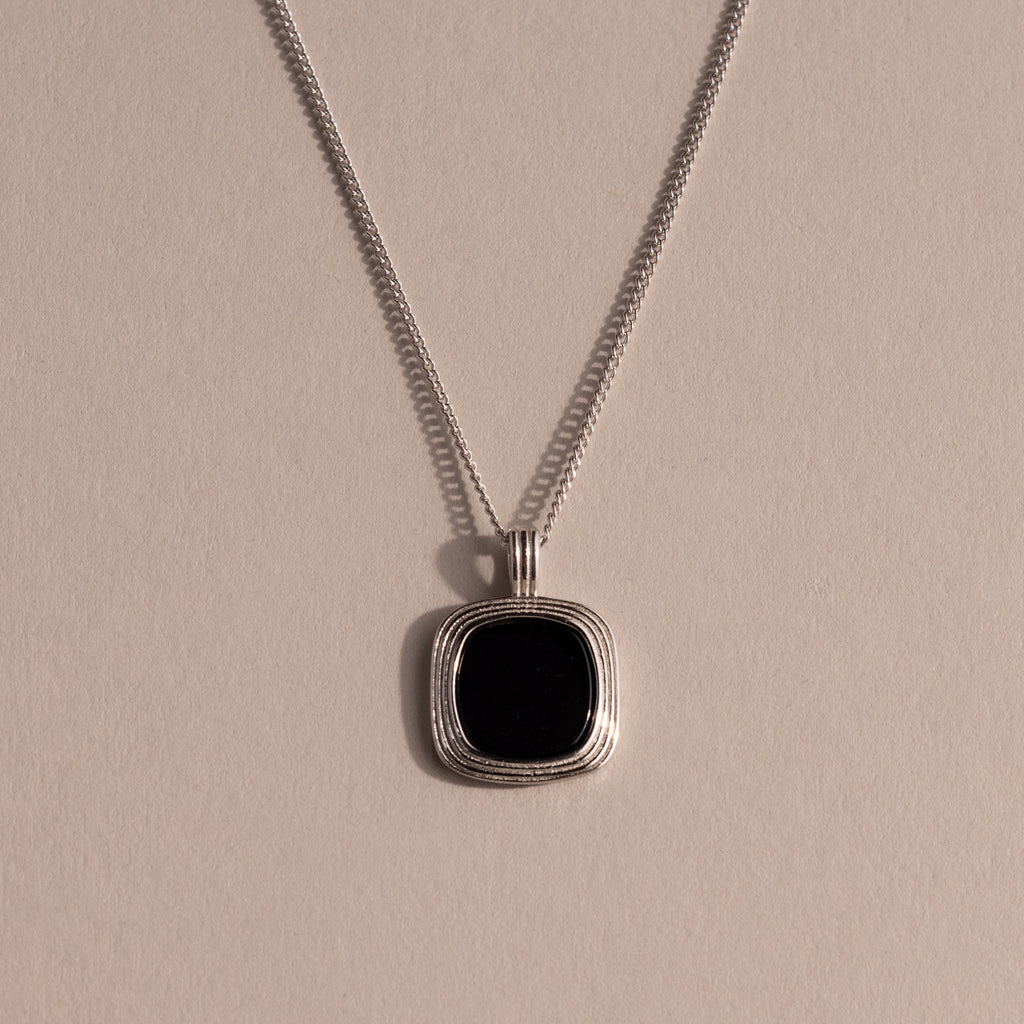 Silver chain necklace featuring a square black onyx pendant in a textured gold bezel, displayed against a neutral background with soft lighting.