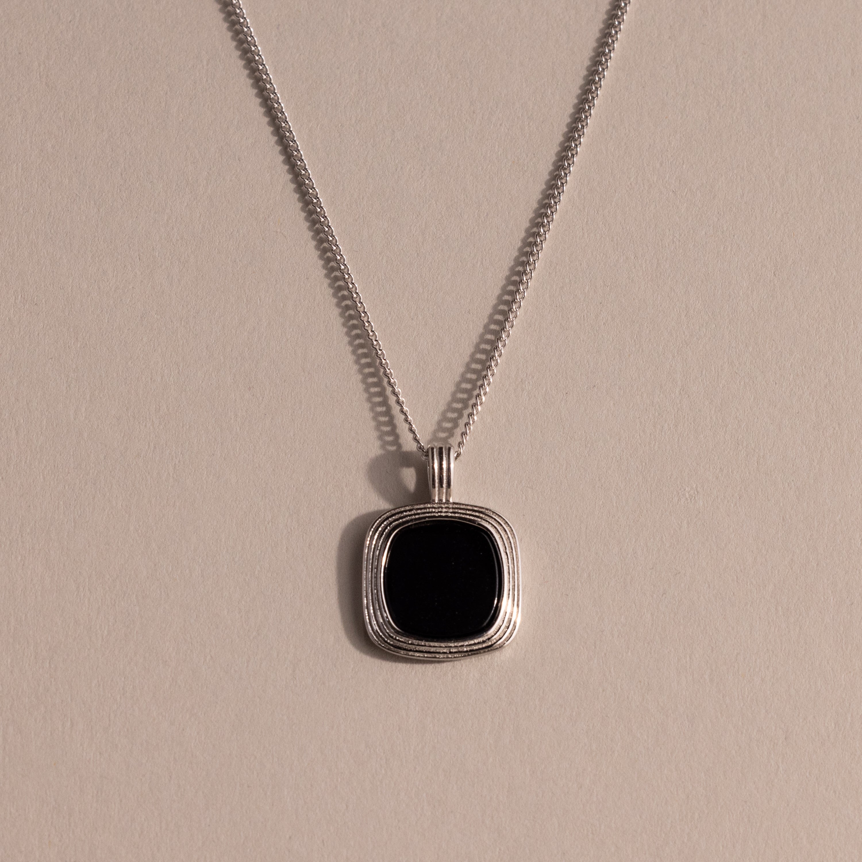 Silver chain necklace featuring a square black onyx pendant in a textured gold bezel, displayed against a neutral background with soft lighting.
