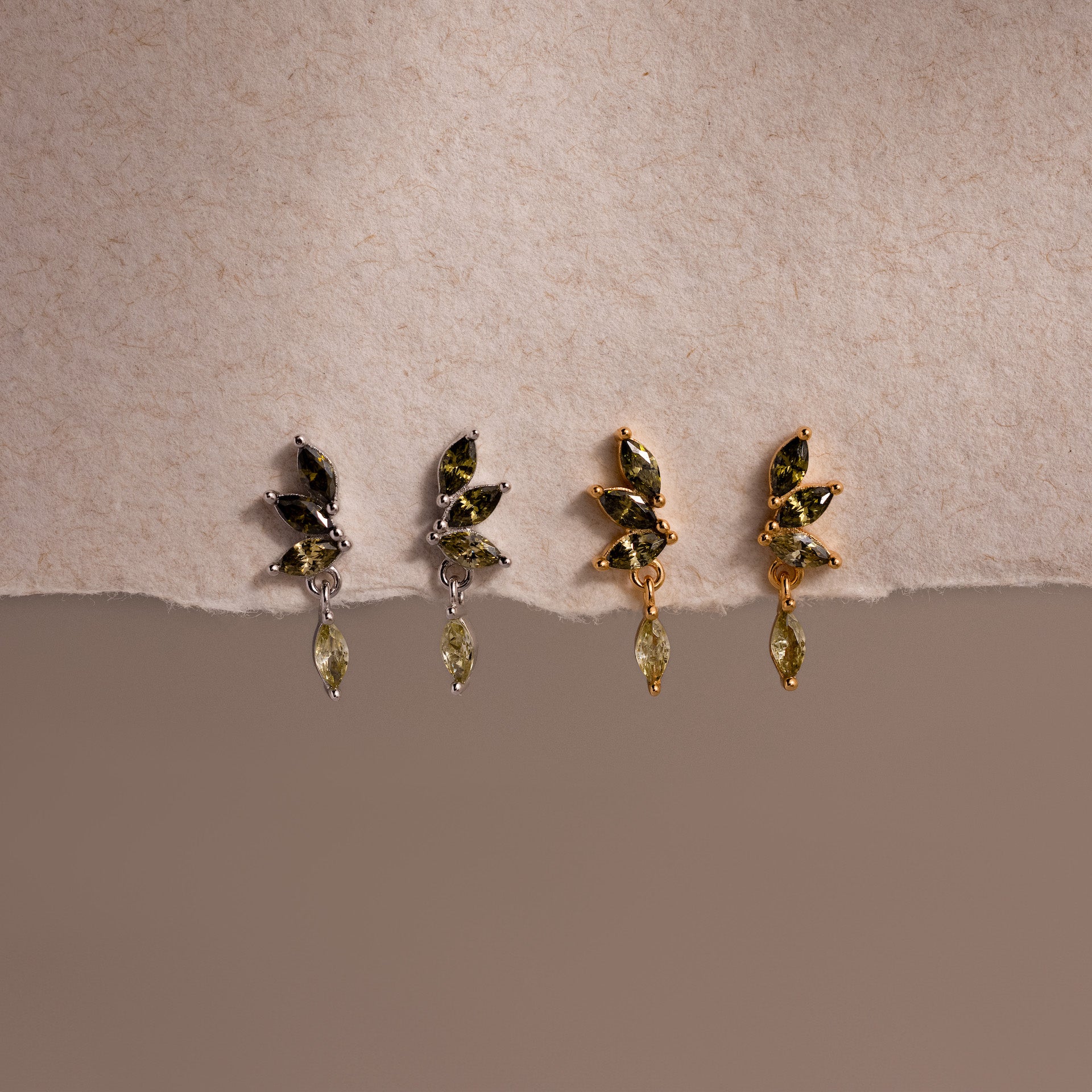 Two pairs of marquise-cut drop earrings in gold and silver, each with green peridot gemstones arranged in a cascading leaf-inspired design, displayed on a soft cream fabric.