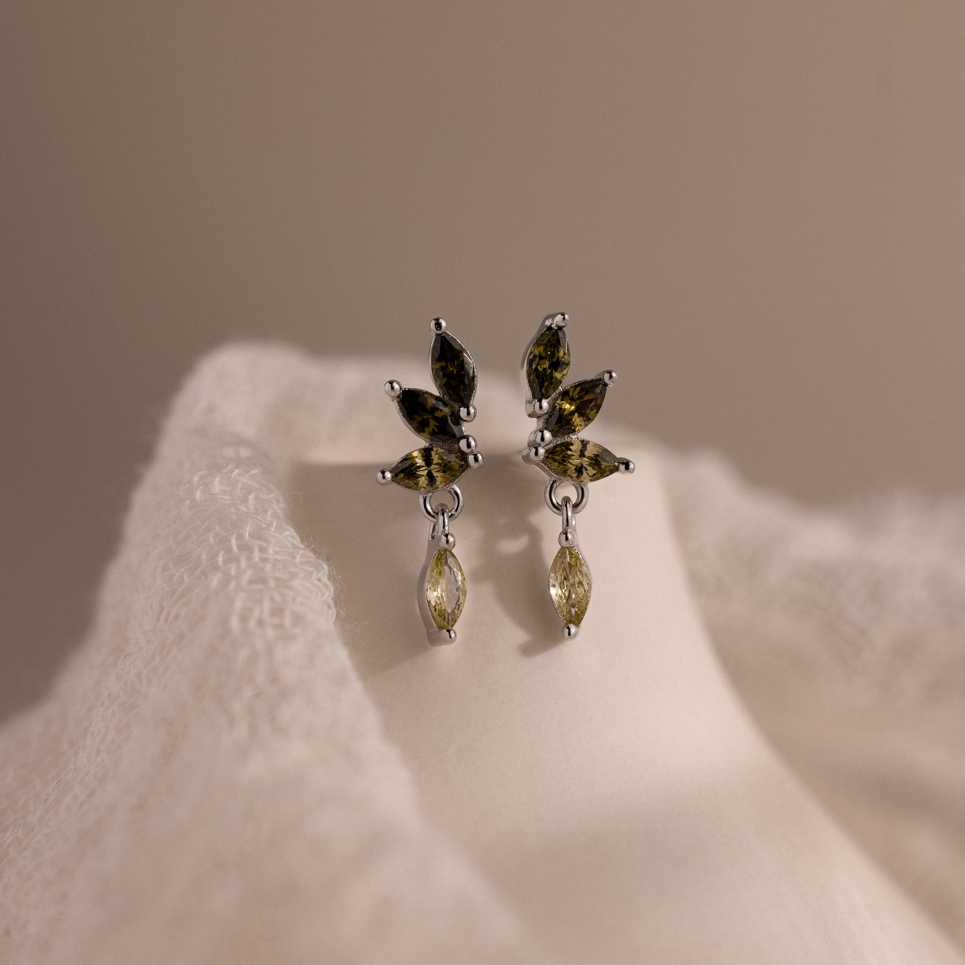 Silver marquise-cut drop earrings with green peridot gemstones arranged in a cascading leaf-inspired design, displayed on a white fabric.