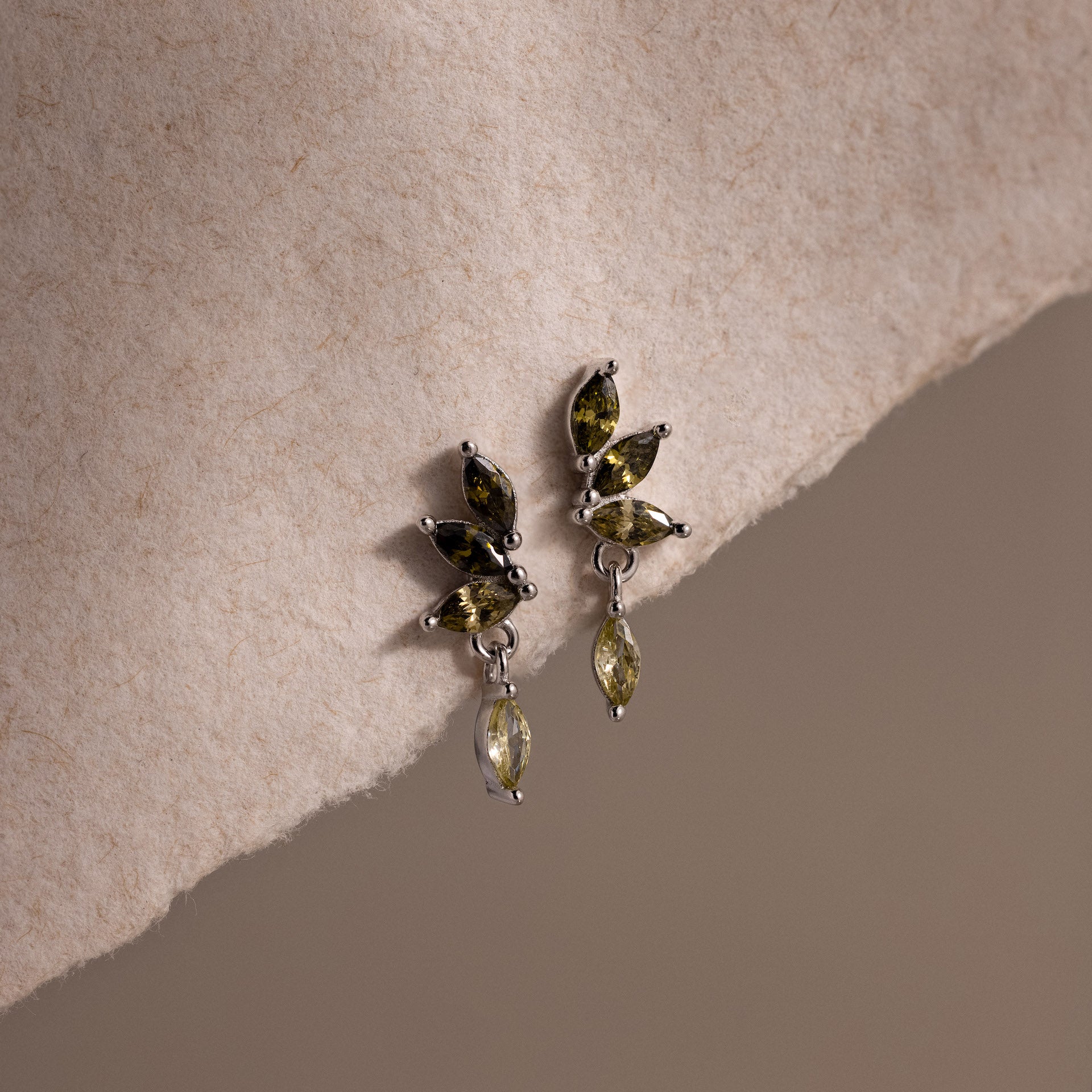 Silver marquise-cut drop earrings with green peridot gemstones arranged in a cascading leaf-inspired design, displayed on a neutral fabric.