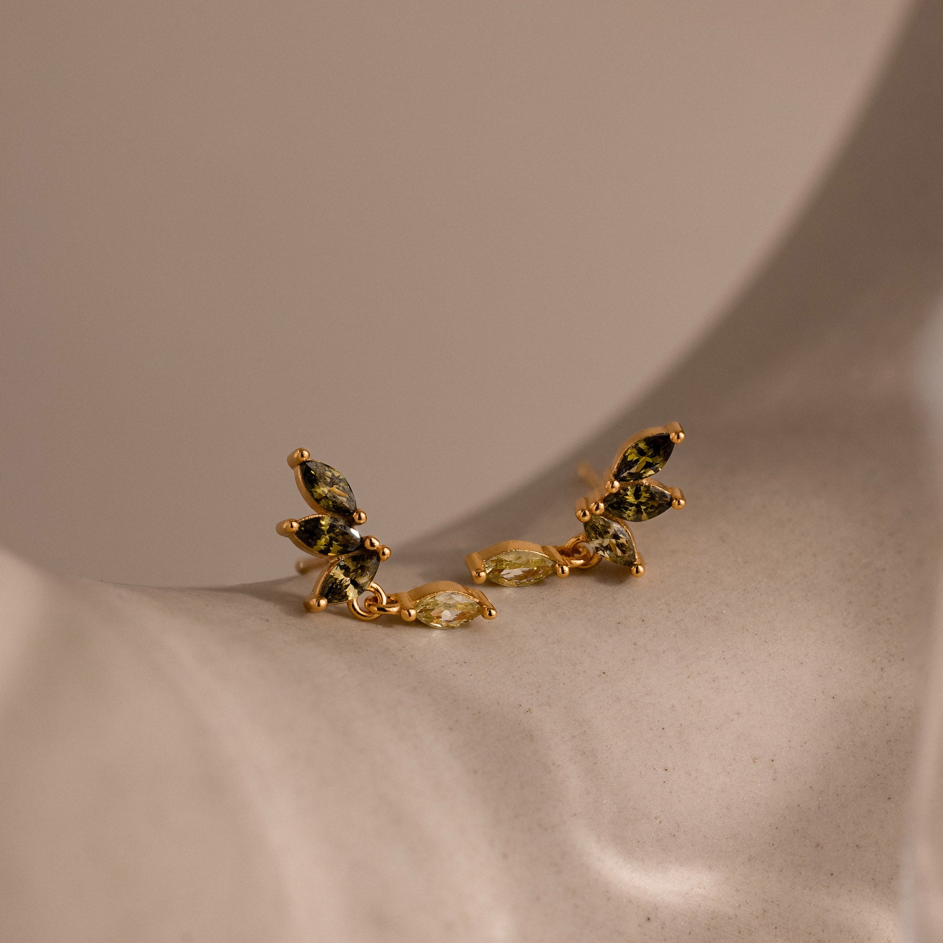 Gold marquise-cut drop earrings with green peridot gemstones arranged in a cascading leaf-inspired design, displayed on a soft ceramic surface.