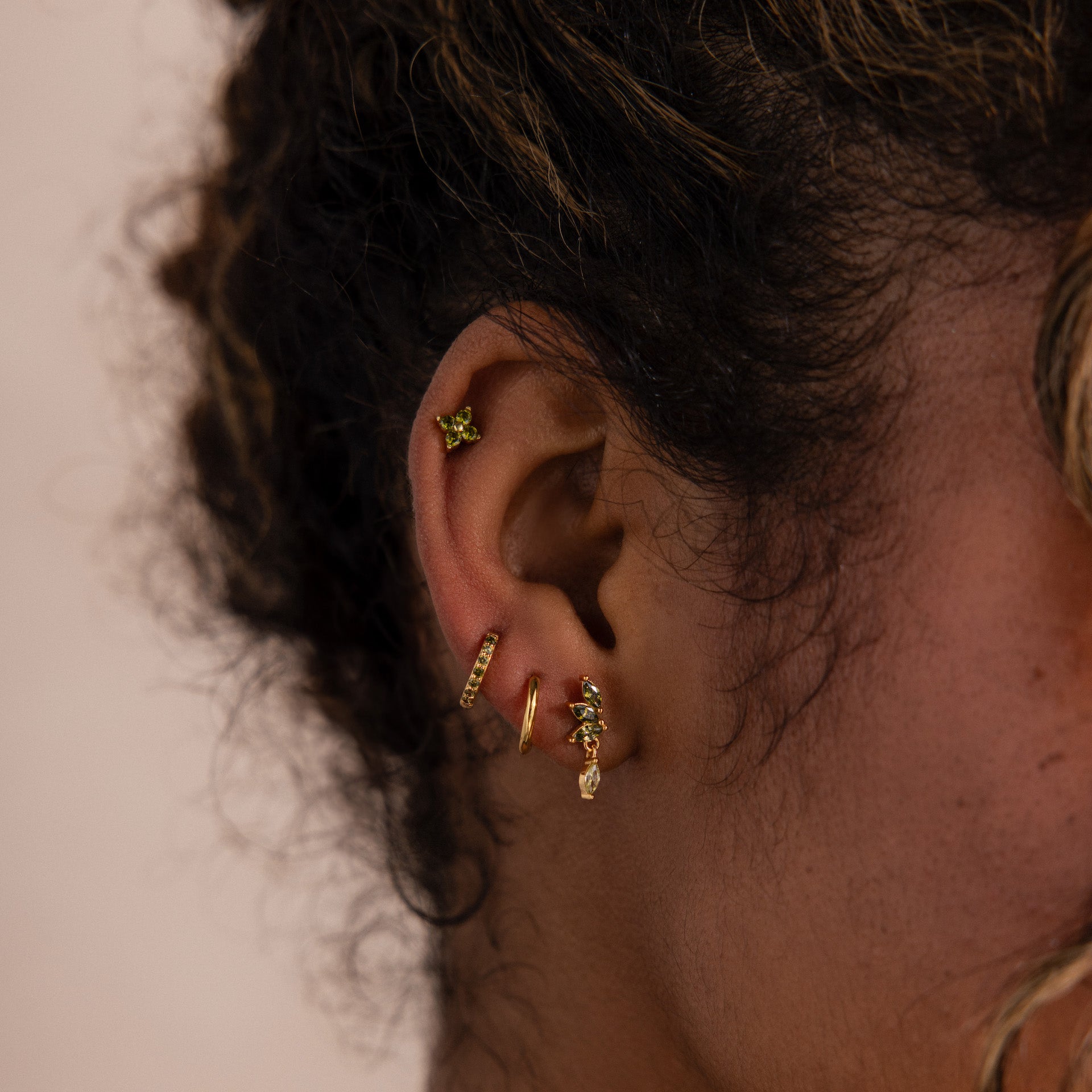 Model wearing a gold marquise-cut drop stud earring with green peridot gemstones, stacked with two matching huggie hoop earrings and one stud earring.