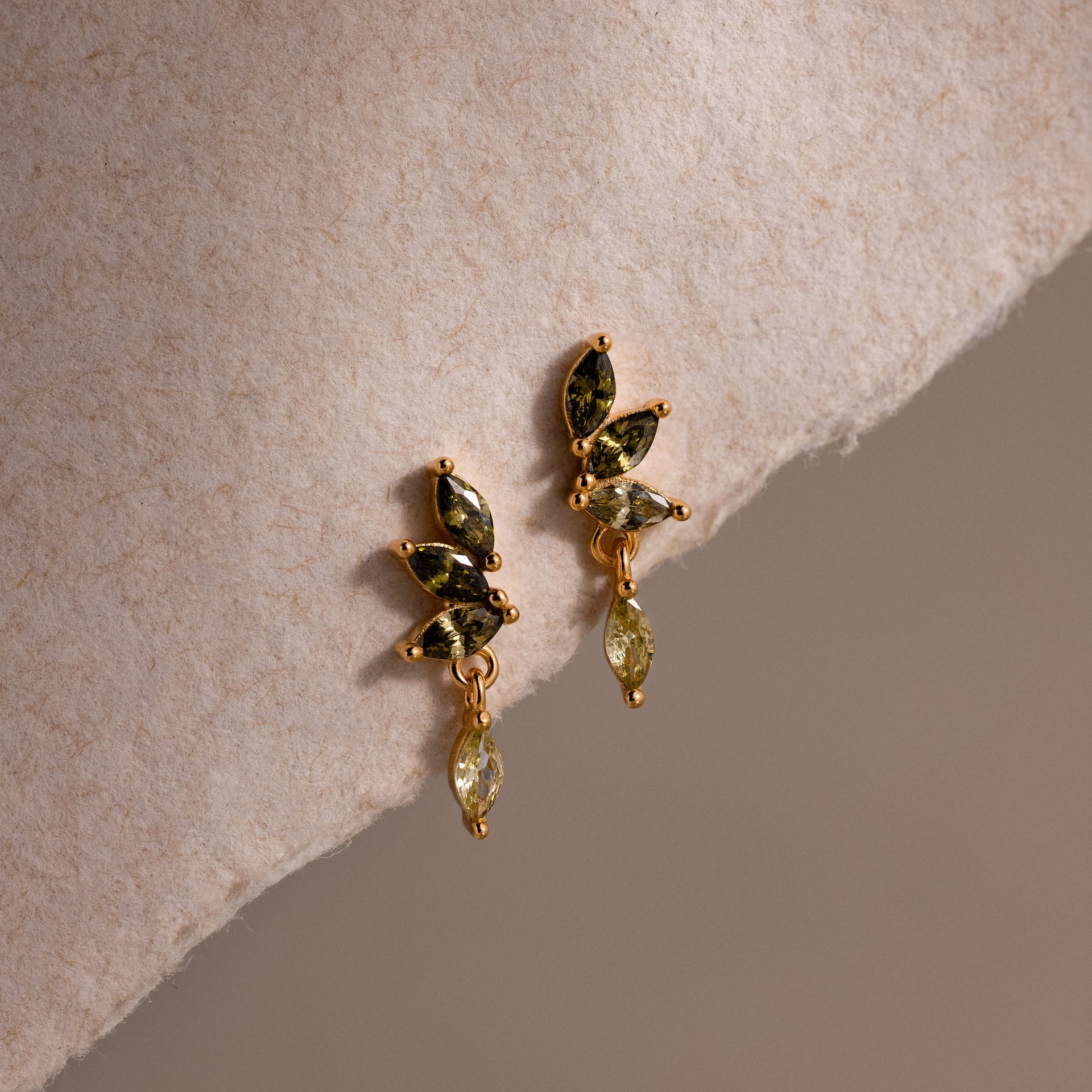 Gold marquise-cut drop earrings with green peridot gemstones arranged in a cascading leaf-inspired design, displayed on a soft cream fabric.