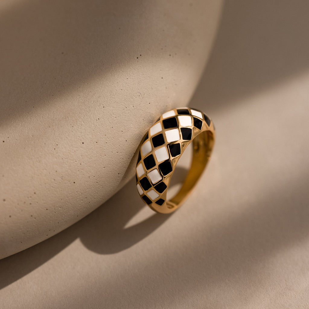 Gold dome ring with black and white checkered enamel pattern, displayed up-right against a neutral surface.
