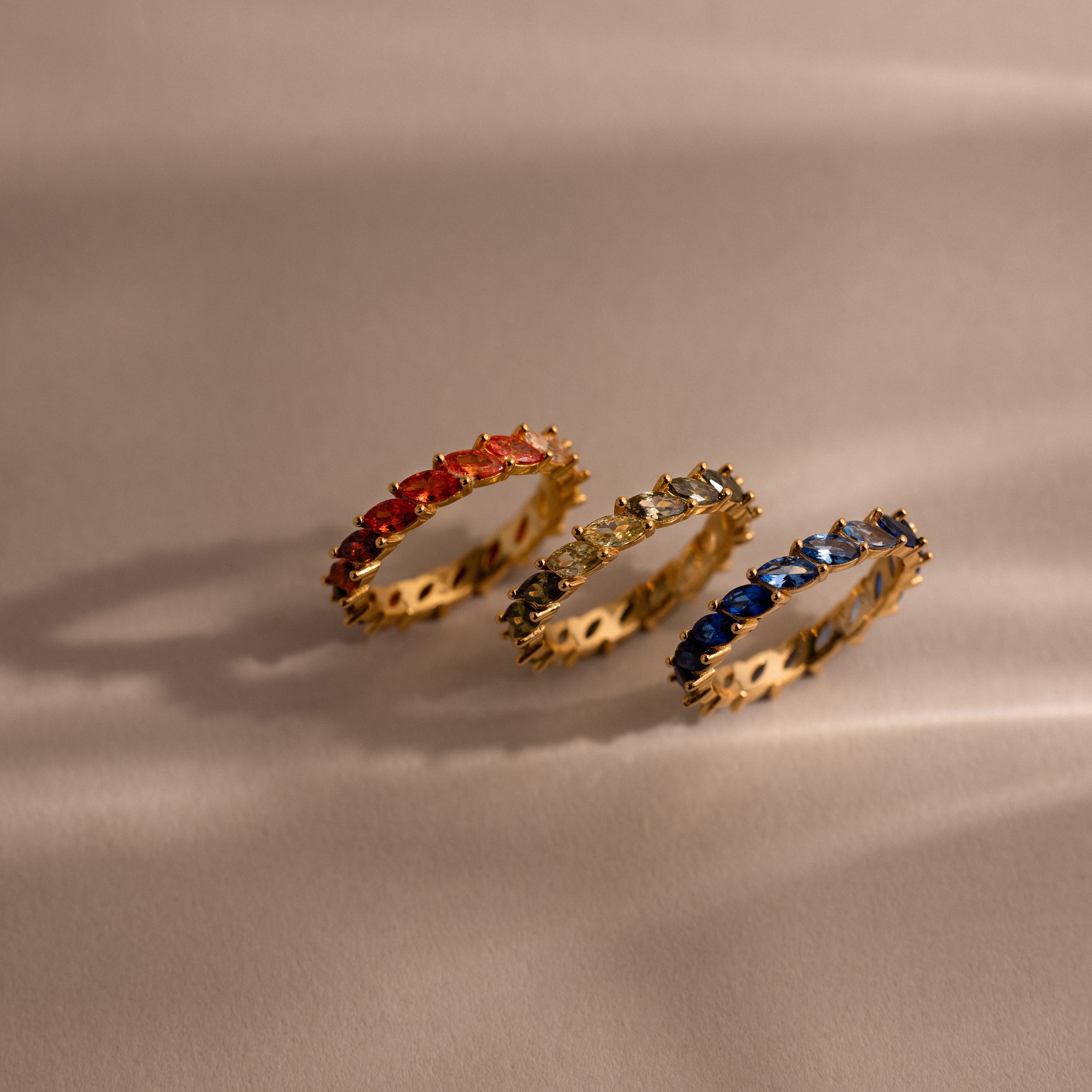 Three gold rings with colorful red, green and blue ombre gemstones on a beige fabric background.