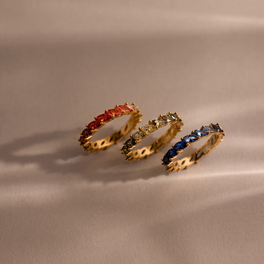 Three gold rings with colorful red, green and blue ombre gemstones on a beige fabric background.