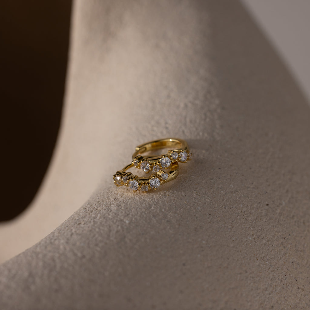 Pair of Marquise Cluster Hoop Earrings in gold with clustered CZ diamond stones, displayed sideways on a neutral stone surface.