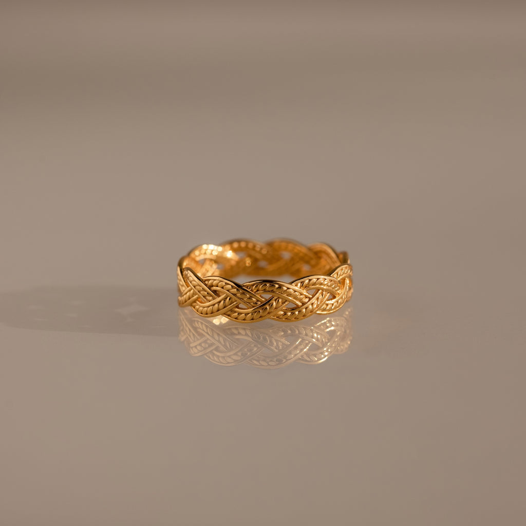 The Woven Ring features a gold braided silhouette and intricate woven design, showcased on a reflective, neutral surface.