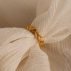 The Woven Ring, featuring a gold braided design, rests on soft cream fabric with delicate folds.