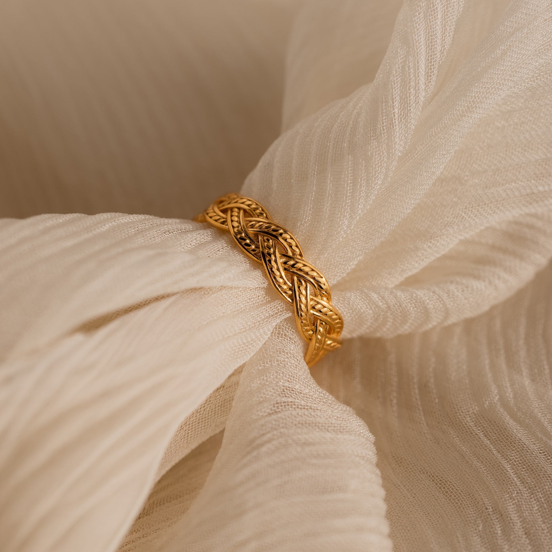 The Woven Ring, featuring a gold braided design, rests on soft cream fabric with delicate folds.