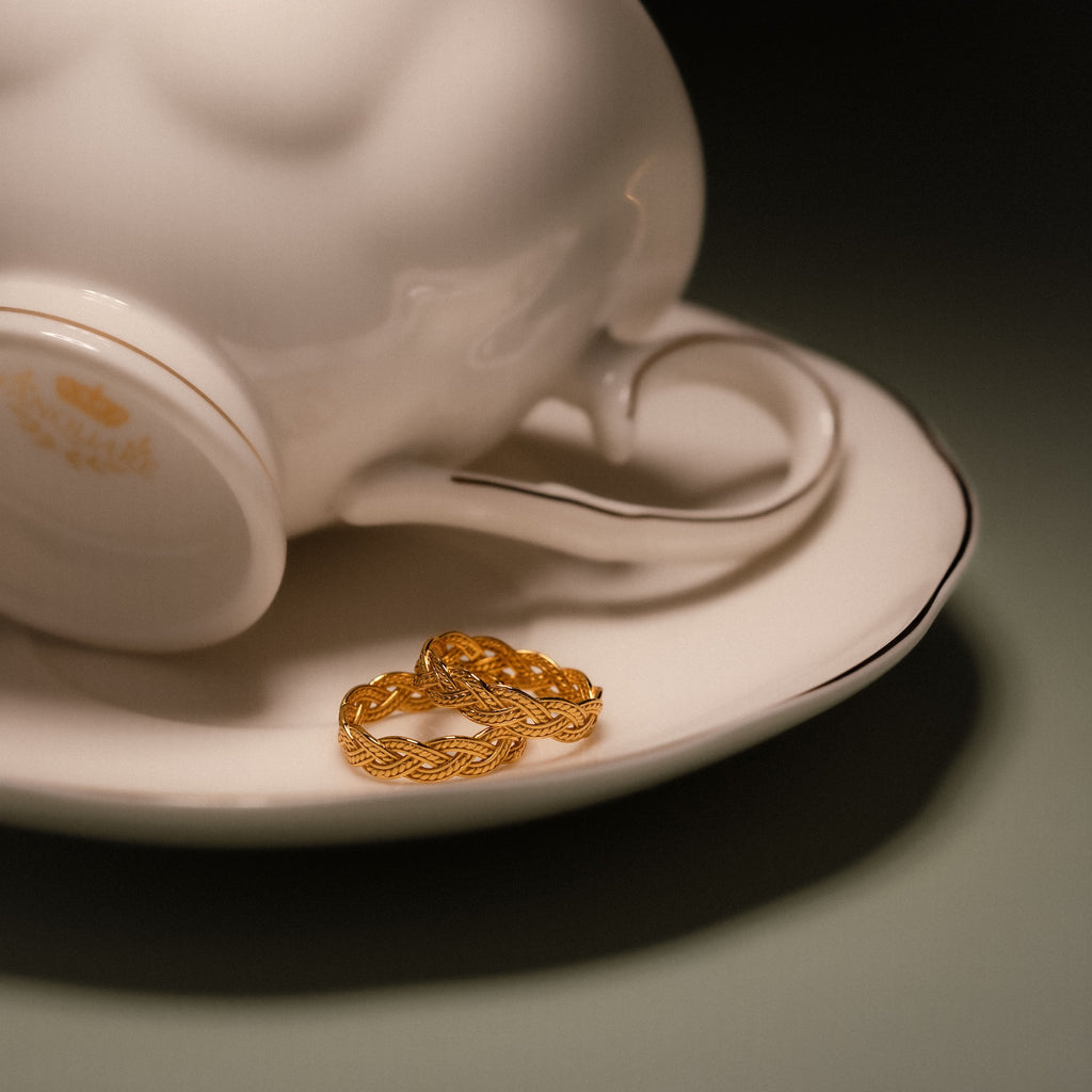 The Woven Ring, featuring a braided gold band, sits on a white saucer next to an upside-down teacup adorned with a subtle crest design.