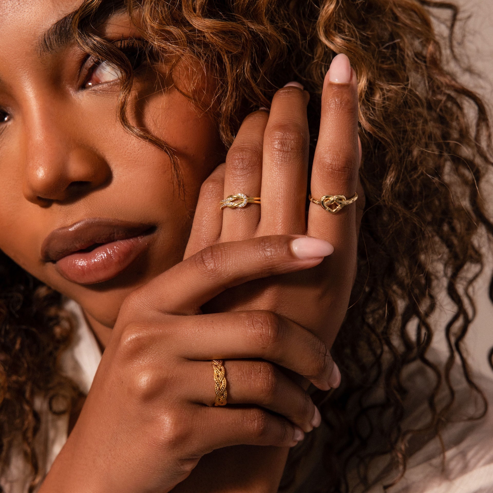 A woman with curly hair shows off a gold ring and the Starry Love Knot Ring on her fingers, gazing at the camera with a neutral expression.