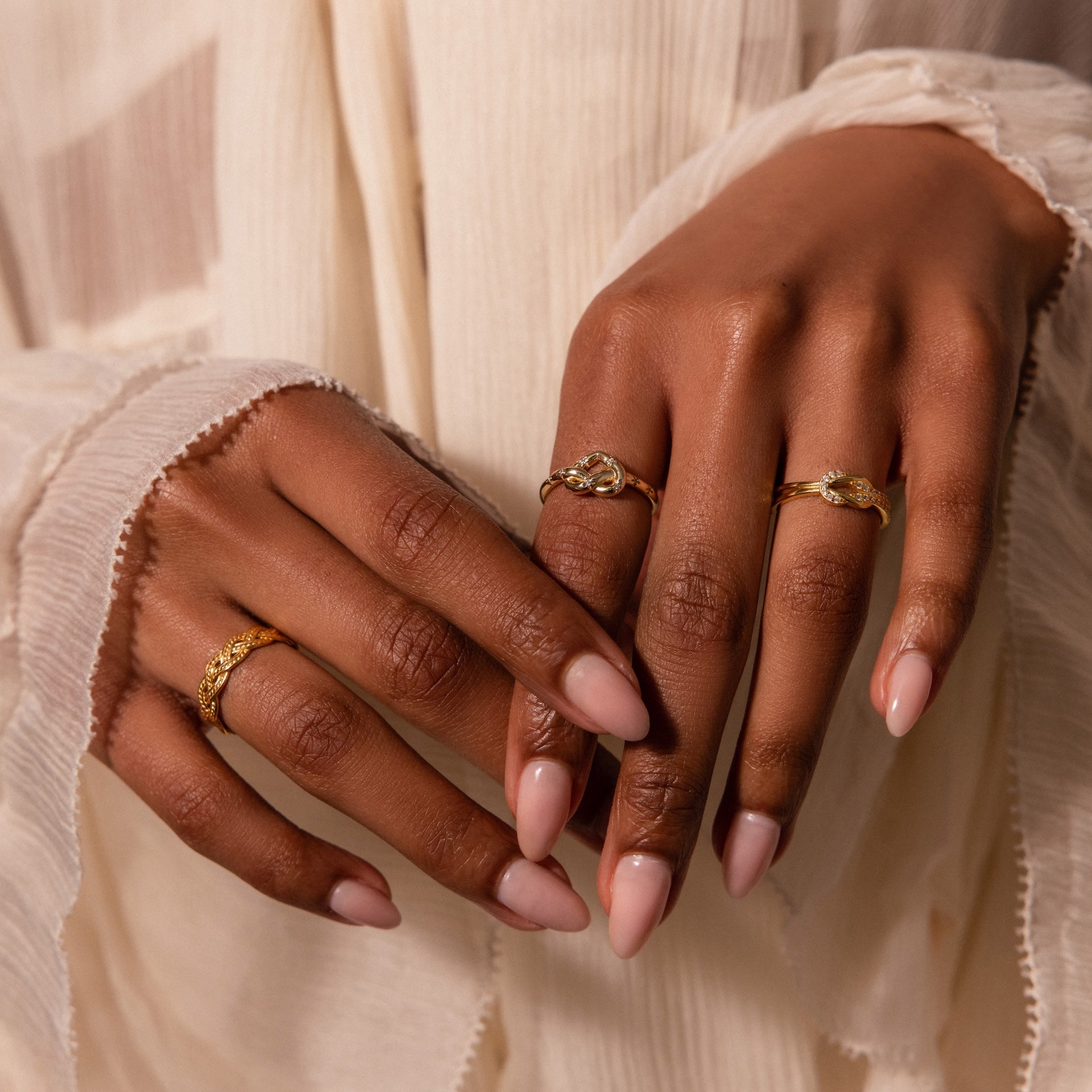 Hands with nude polished nails wear the Starry Love Knot Ring and two gold rings, set against a sheer, cream-colored fabric background.