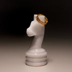 The Woven Ring, featuring a braided gold design, is displayed on a white marble chess knight against a dark background.