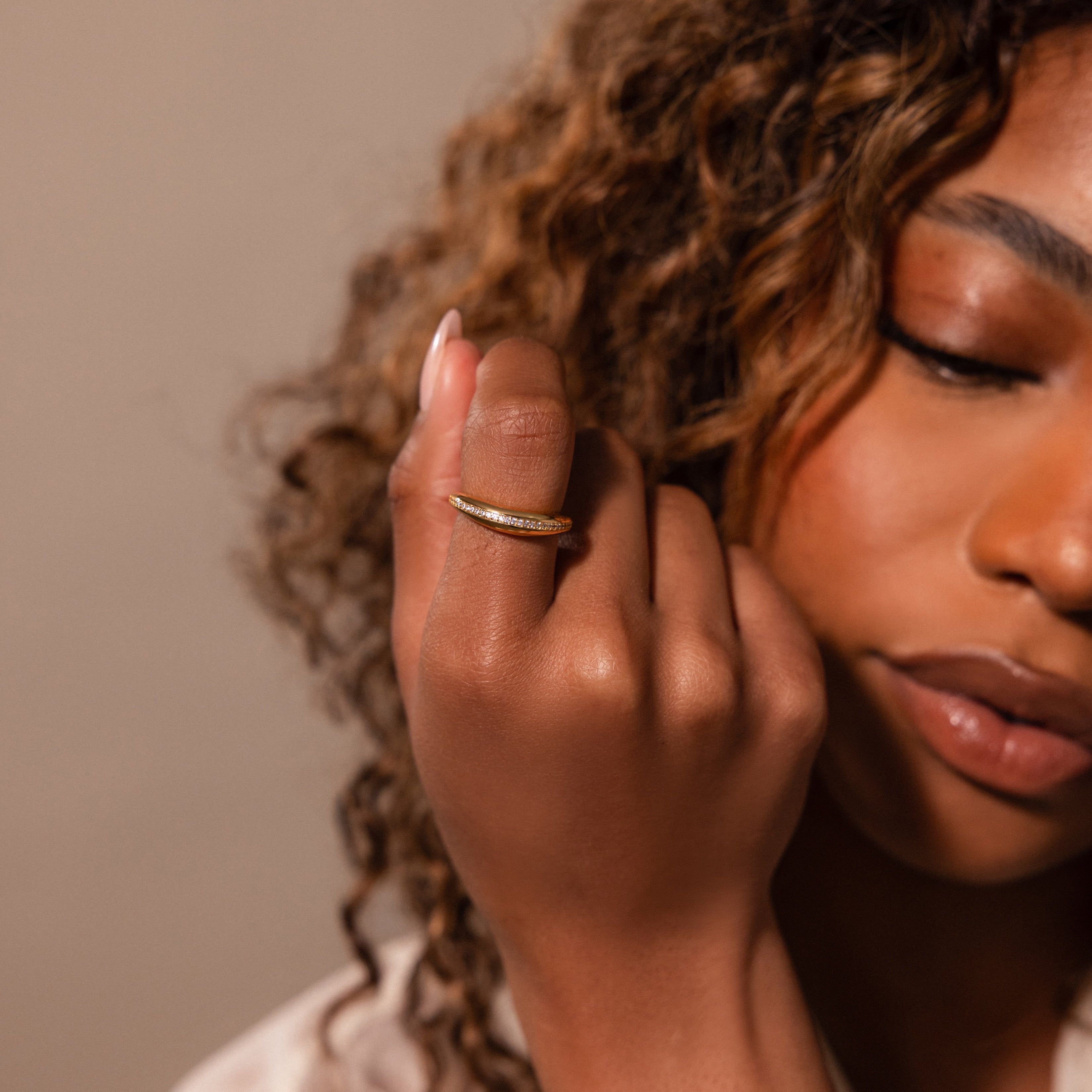 A woman with curly hair wears the Pave Curve Dome Ring on her finger, her face partially visible and eyes closed—an elegant touch to elevate any everyday jewelry stack.
