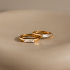 A pair of gold rings, featuring a Diamond Baguette Signet Ring with rectangular clear stones, are displayed elegantly on a smooth, light-colored surface.