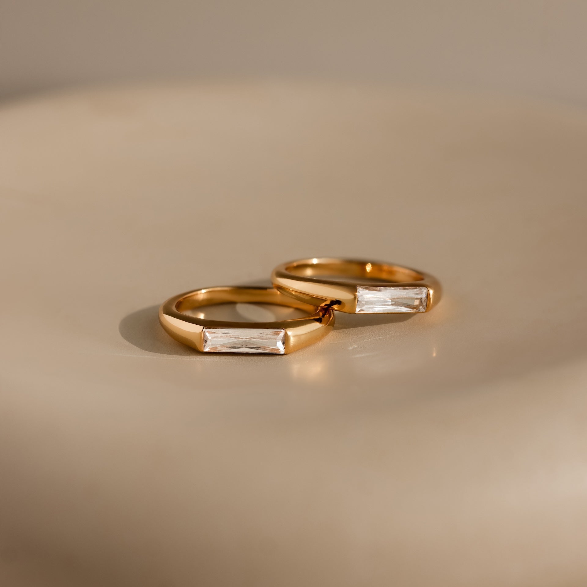 A pair of gold rings, featuring a Diamond Baguette Signet Ring with rectangular clear stones, are displayed elegantly on a smooth, light-colored surface.