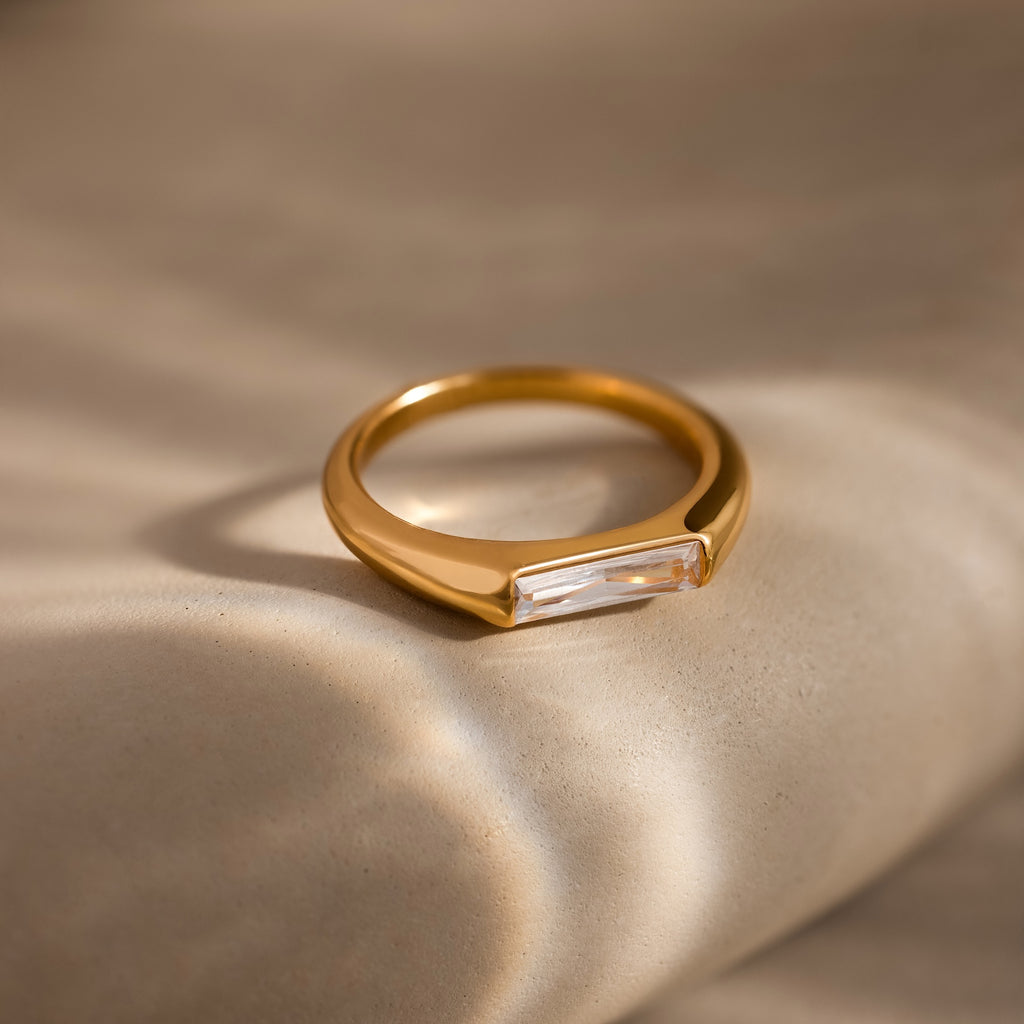 A modern statement piece, the Diamond Baguette Signet Ring showcases a rectangular diamond set in gold, displayed elegantly on a smooth beige surface under soft lighting.