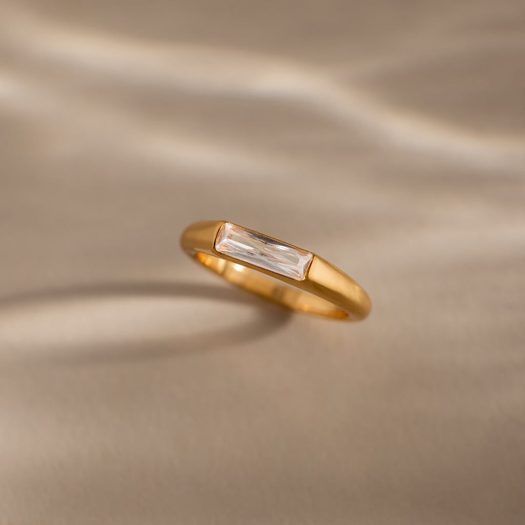 A modern statement piece, the Diamond Baguette Signet Ring showcases a rectangular clear diamond, elegantly displayed on a beige surface with soft light and shadows.