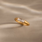 A modern statement piece, the Diamond Baguette Signet Ring showcases a rectangular clear diamond, elegantly displayed on a beige surface with soft light and shadows.