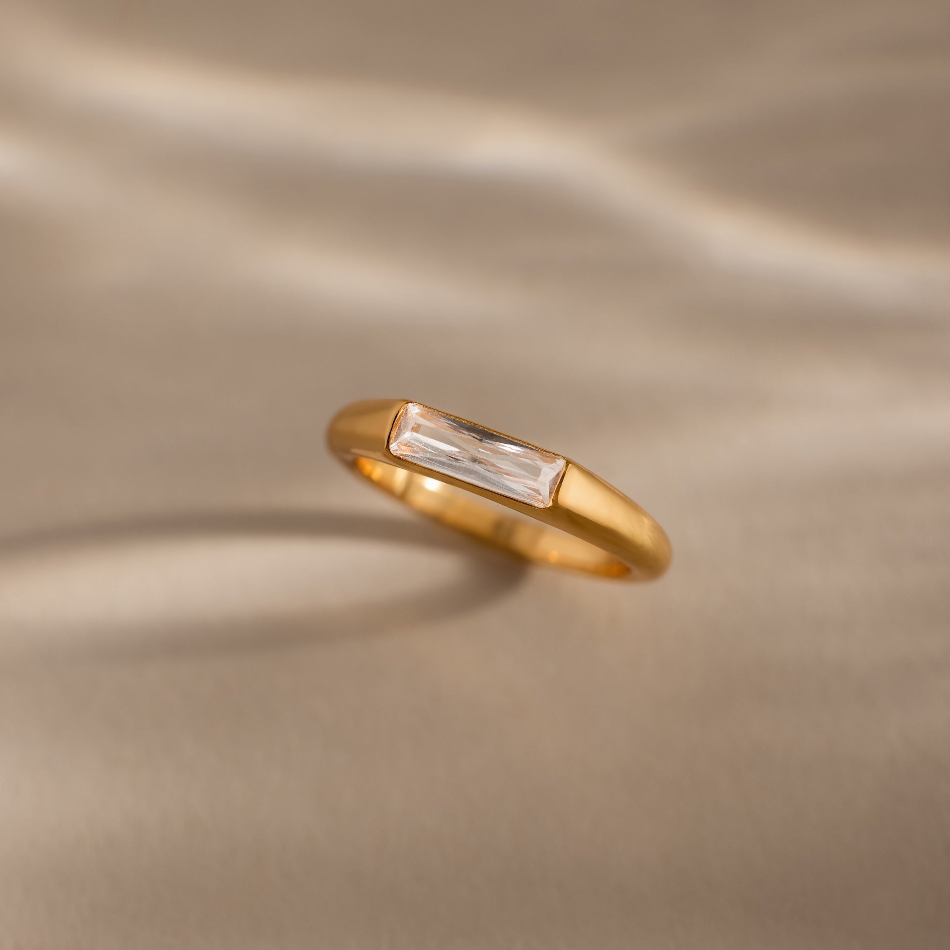 A modern statement piece, the Diamond Baguette Signet Ring showcases a rectangular clear diamond, elegantly displayed on a beige surface with soft light and shadows.