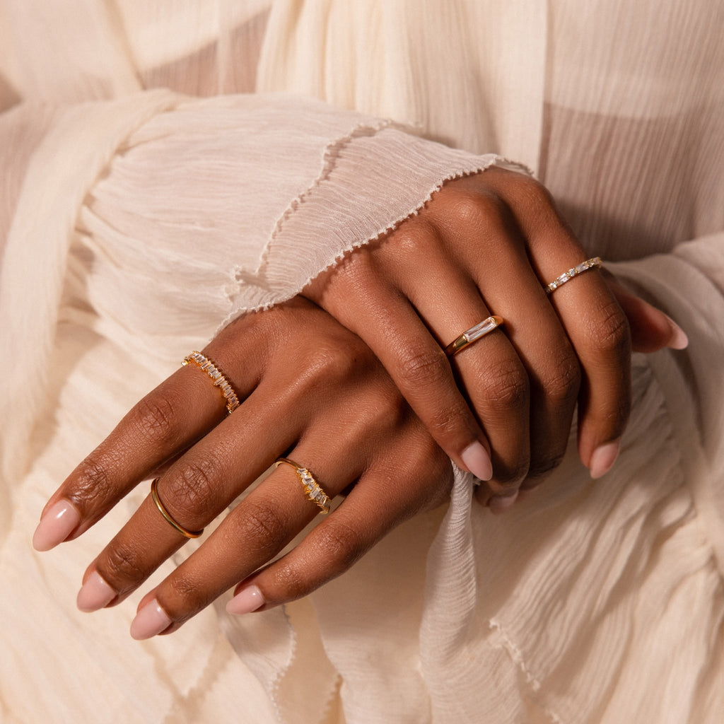 Two hands with manicured nails are adorned with delicate gold rings, featuring the modern Diamond Baguette Signet Ring, shown against cream-colored pleated fabric.