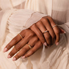 Two hands with manicured nails are adorned with delicate gold rings, featuring the modern Diamond Baguette Signet Ring, shown against cream-colored pleated fabric.