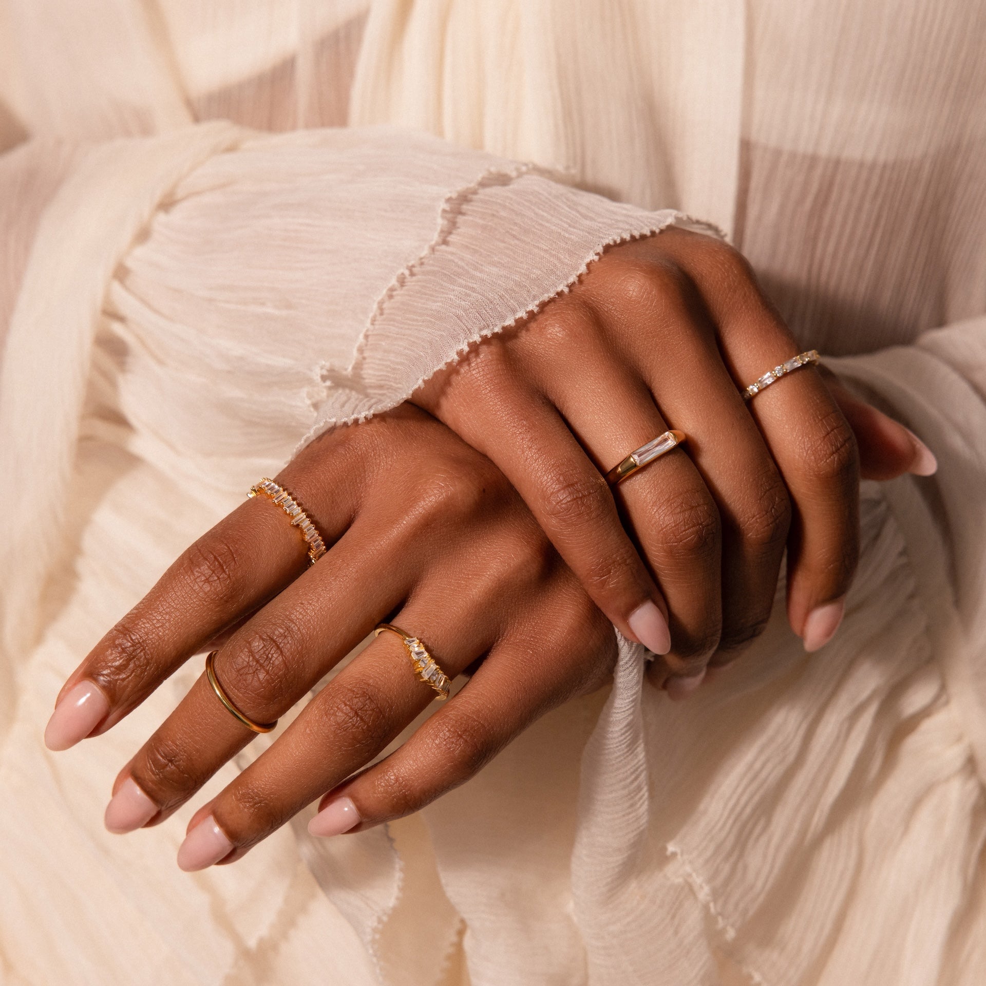 Hands with nude nails showcasing delicate gold and diamond rings, featuring the Baguette Signet Ring, against a cream pleated fabric background.