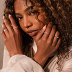 Woman with curly hair wears the Diamond Baguette Signet Ring with gold bands and a light, textured blouse, posing with hands near her face.