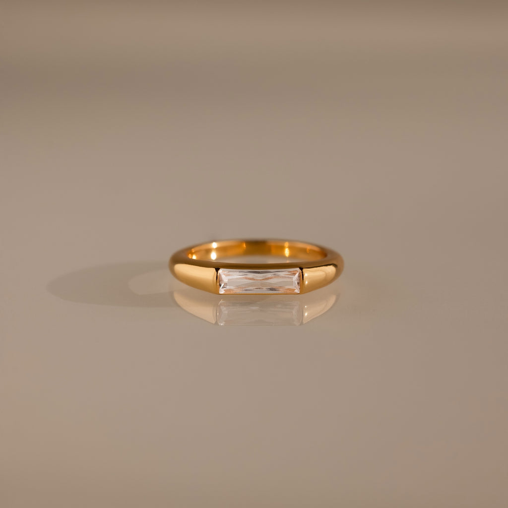 Diamond Baguette Signet Ring: This modern gold ring features a rectangular diamond centerpiece, showcased on a reflective beige surface.