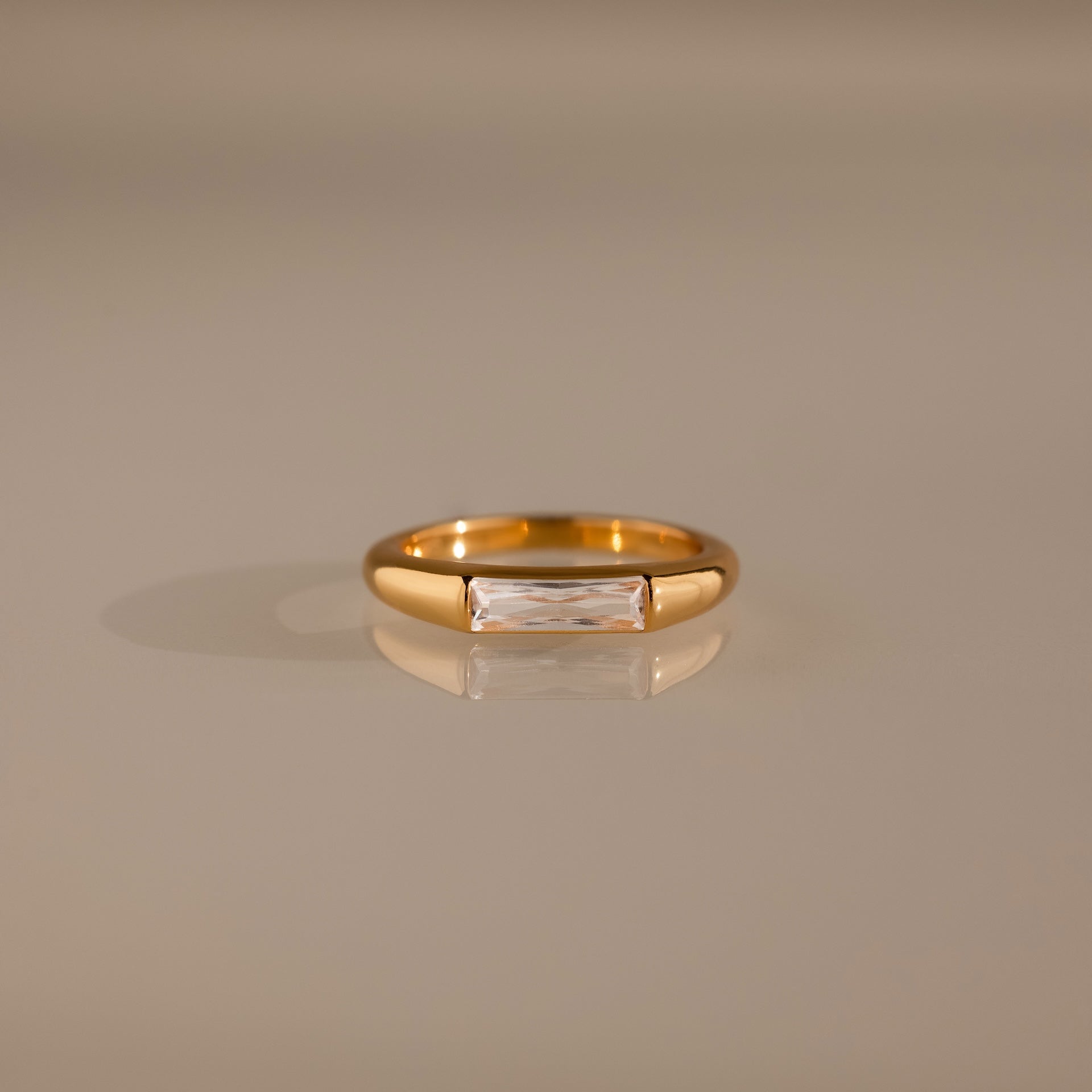 Diamond Baguette Signet Ring: This modern gold ring features a rectangular diamond centerpiece, showcased on a reflective beige surface.