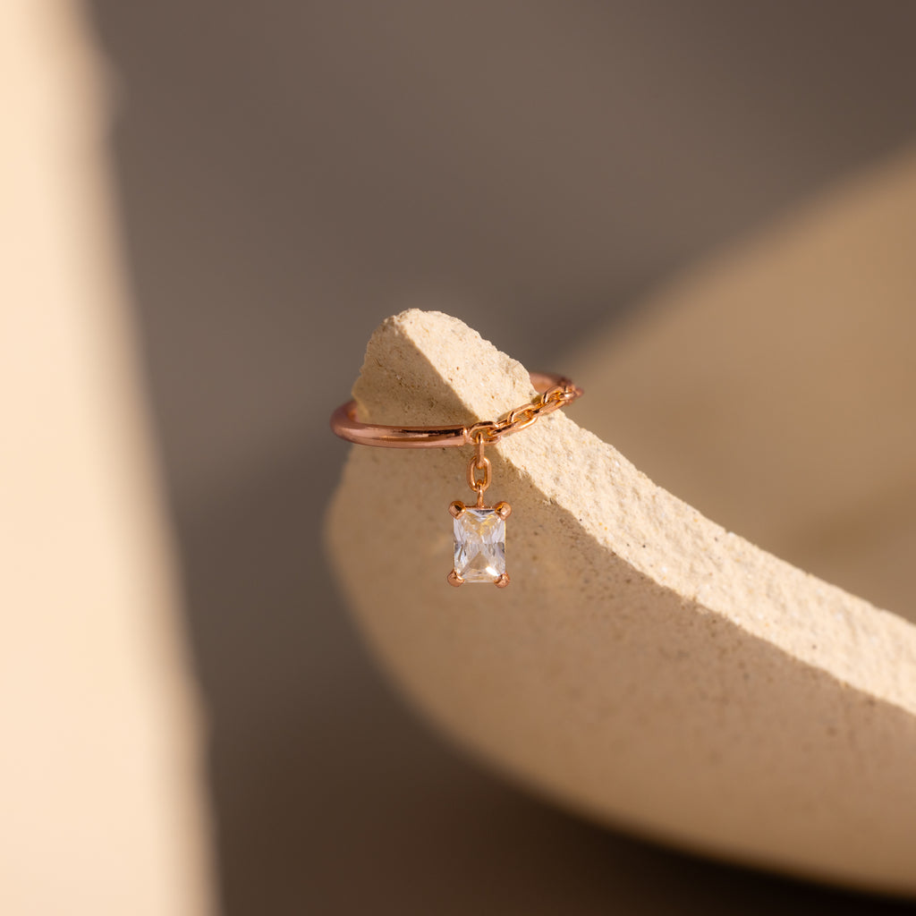 The Diamond Drop Chain Ring, featuring a dazzling diamond on a gold chain, rests on a textured beige surface illuminated by soft light.