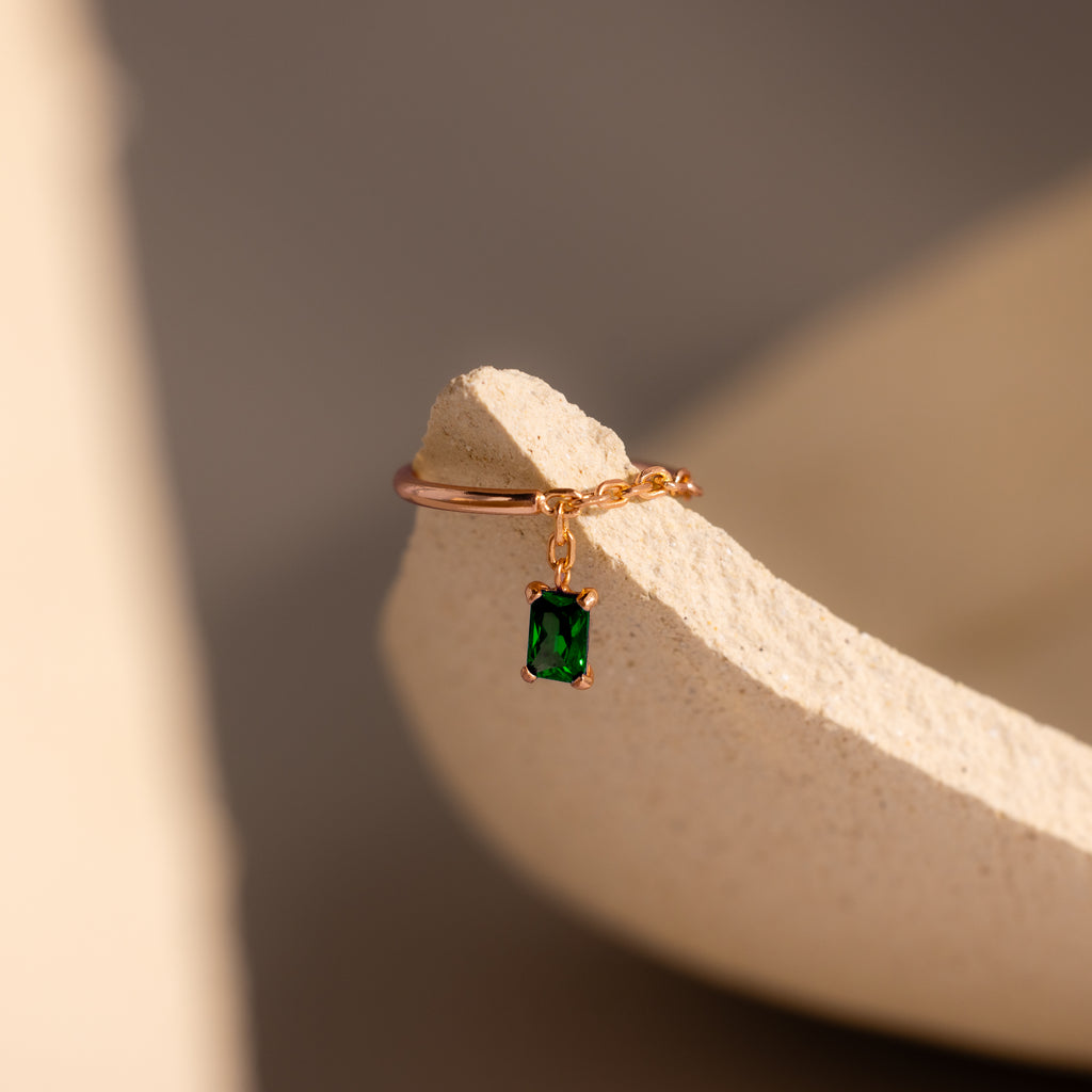 The Emerald Drop Chain Ring features a gold chain band with a dangling emerald-cut green stone, showcased on a textured beige surface.