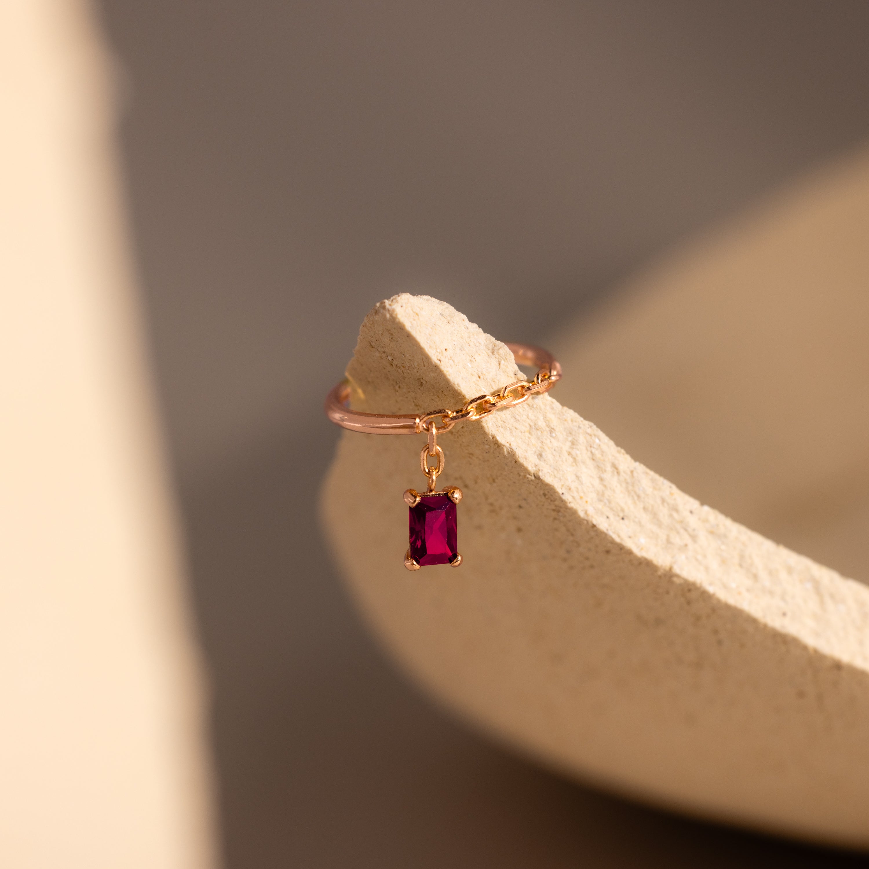 The Ruby Drop Chain Ring features a delicate gold chain and a small rectangular red stone, displayed on a textured beige surface in soft light.