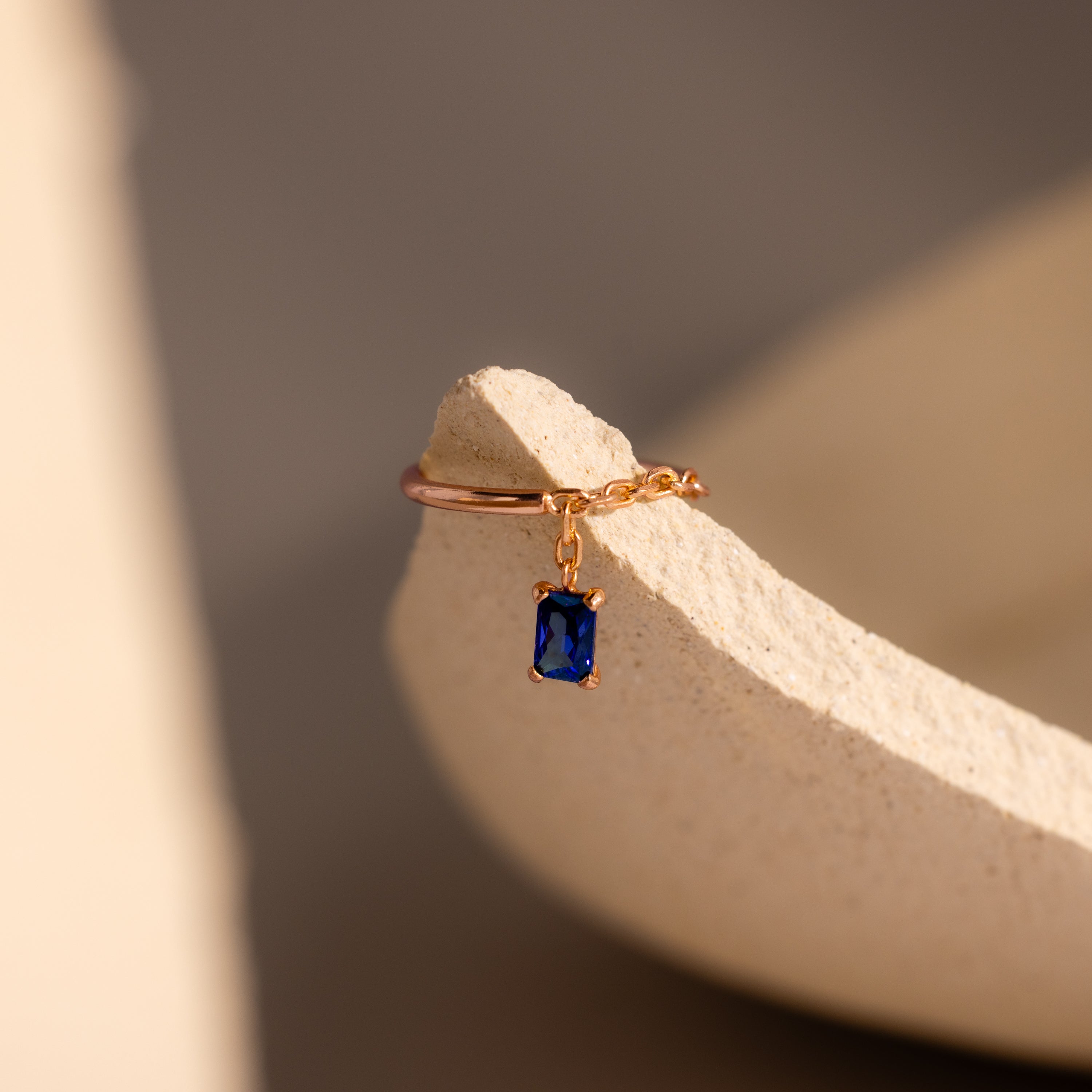 The Sapphire Drop Chain Ring, featuring a dangling blue charm, is displayed on a textured beige ceramic surface.