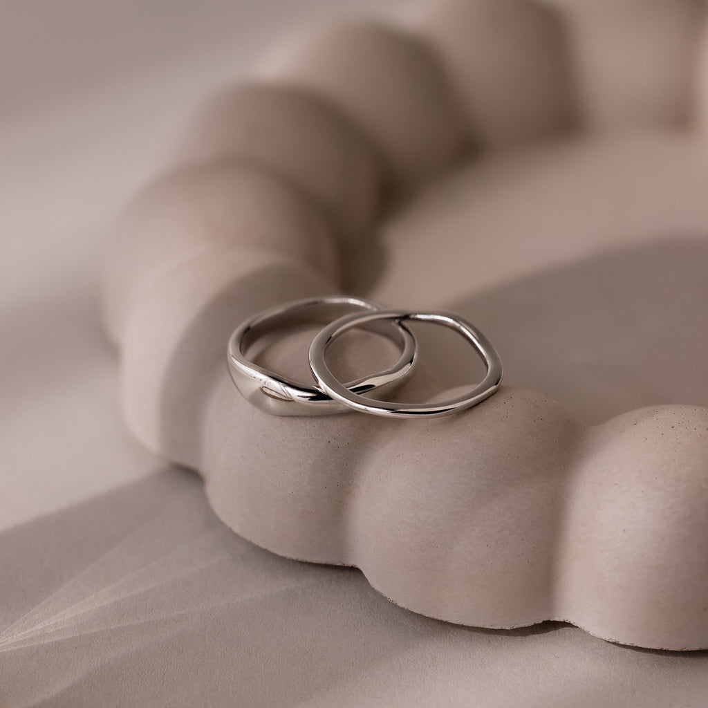 The Duo Form Ring Set, featuring two simple silver rings, rests on a textured, light-toned rounded surface under soft lighting.