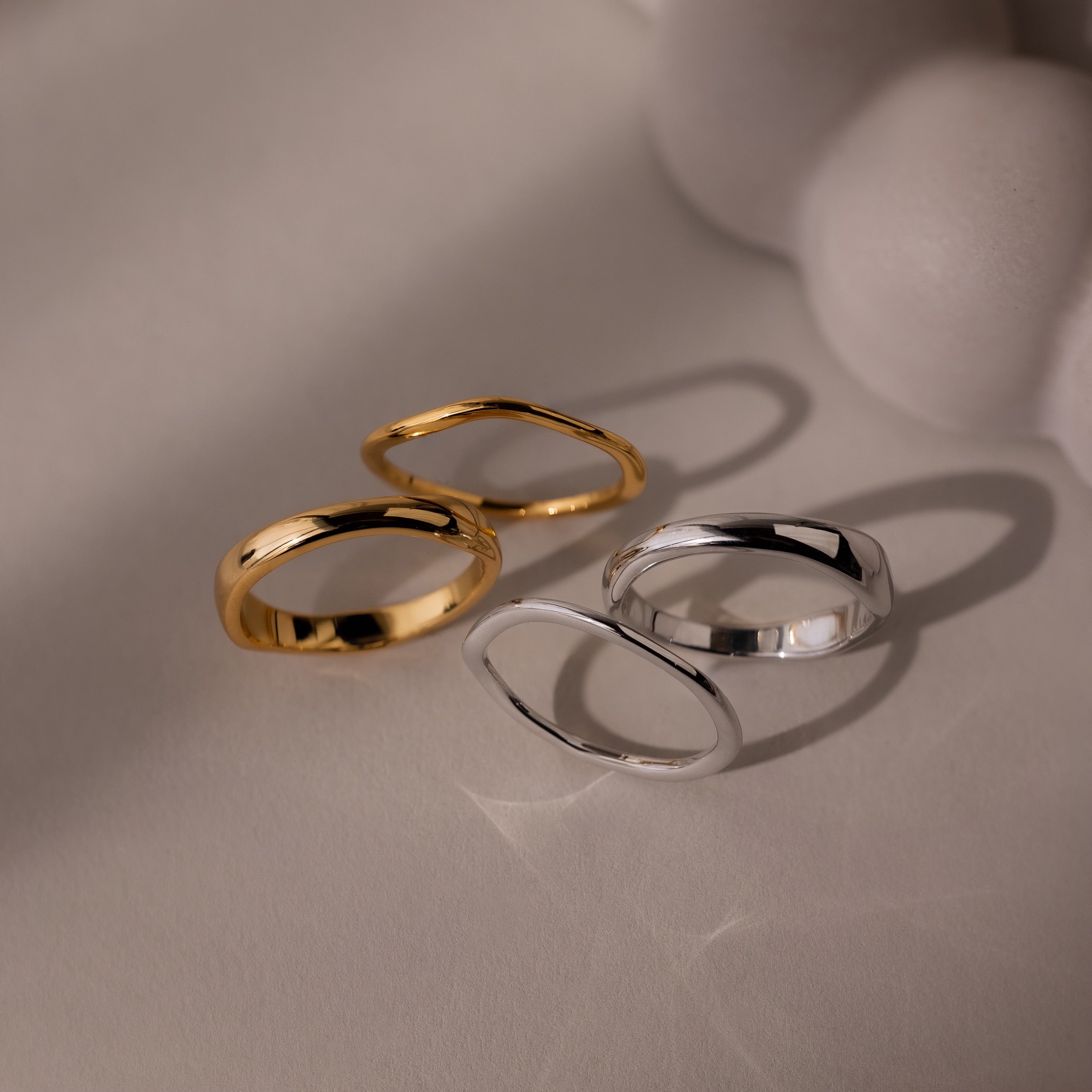 The Duo Form Ring Set, featuring two pairs of gold and silver ring sets with sculptural design, rests on a light surface casting soft shadows beside a blurred white object.