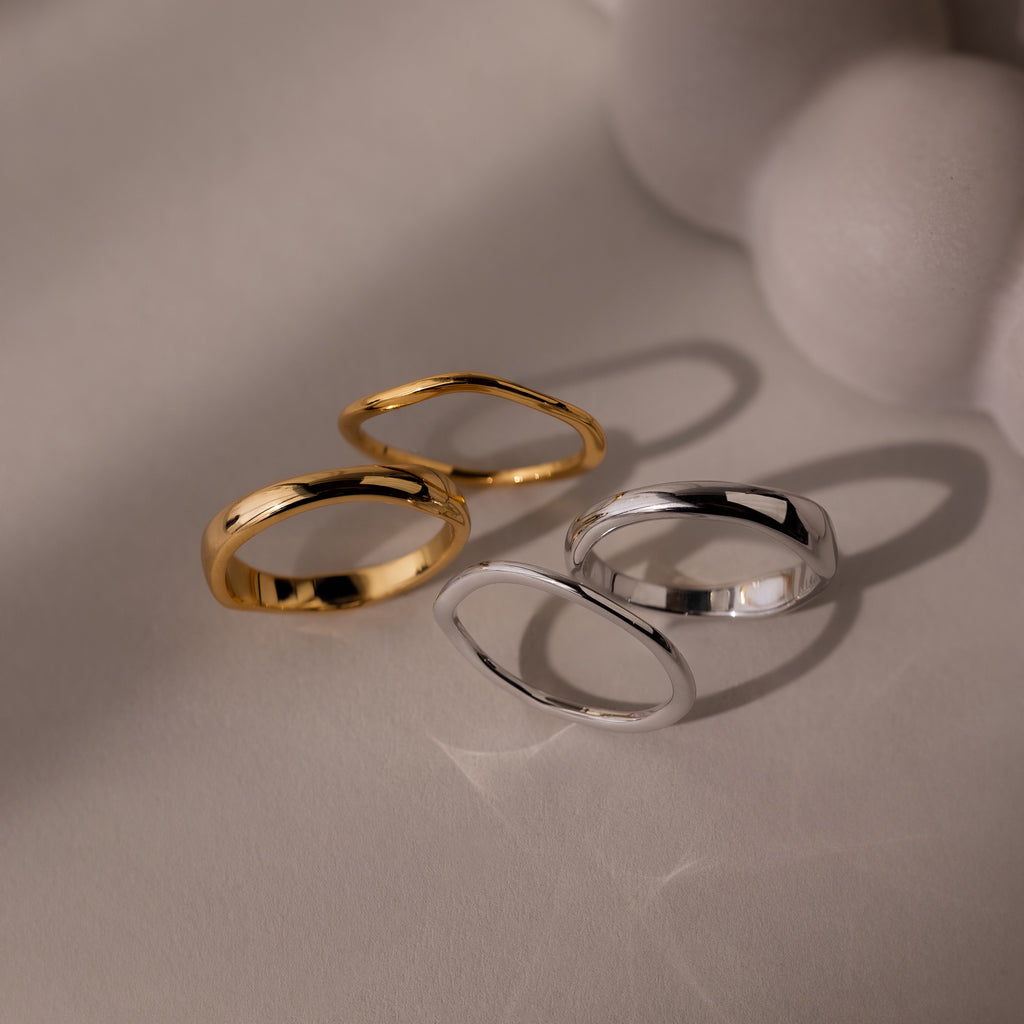 The Duo Form Ring Set, featuring two pairs of gold and silver ring sets with sculptural design, rests on a light surface casting soft shadows beside a blurred white object.
