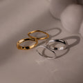 The Duo Form Ring Set, featuring two pairs of gold and silver ring sets with sculptural design, rests on a light surface casting soft shadows beside a blurred white object.
